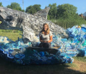botanical garden intern smiling white sitting on a giant shark that is part of a sculpture exhibit where it's made of trash from the ocean