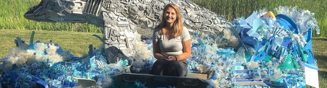 botanical garden intern smiling white sitting on a giant shark that is part of a sculpture exhibit where it's made of trash from the ocean
