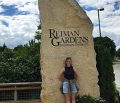 botanical garden intern smiling outside of Reiman Gardens rock in summer with green plants behind it