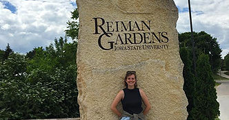 botanical garden intern smiling outside of Reiman Gardens rock in summer with green plants behind it