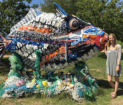 a sculpture piece from a sculpture exhibit that looks like a giant fish with seaweed below it and a person smiling next to it. This sclpture is madee from trash