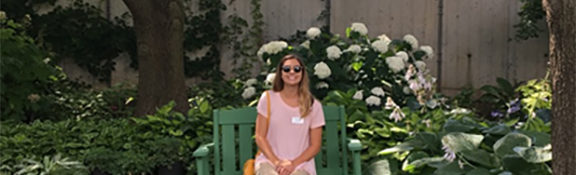 Reiman Gardens intern sitting on a bench and smiling while outside with whtie flwoers behind