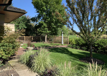 outdoors in summer in the Front Yard Garden with grasses, large trees, a fence, and a building created with the prairie style of architecture