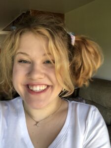 A young woman smiles into the camera. She wears pigtails and looks very happy.